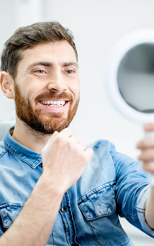 Male dental patient in denim jacket holding mirror and checking smile